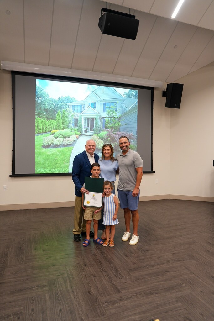 A family with two young children accepts their Westlake in Bloom certificate from the Mayor, their landscaped front yard projected on the screen behind them.
