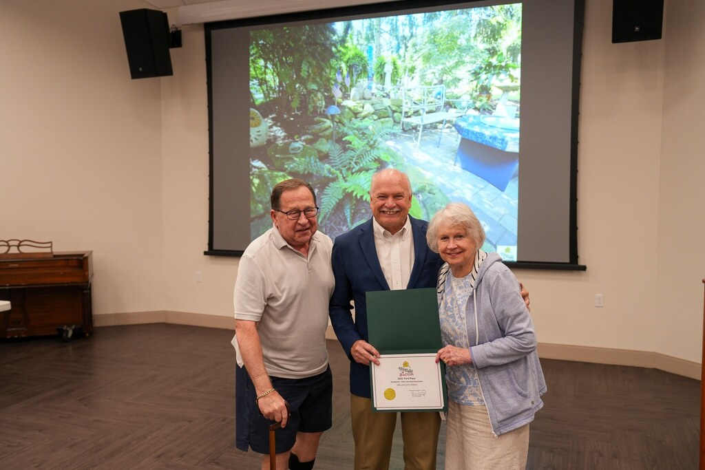 Two Westlake in Bloom recipients stand with the Mayor holding their award certificate, a shaded green garden shown on the screen behind them.