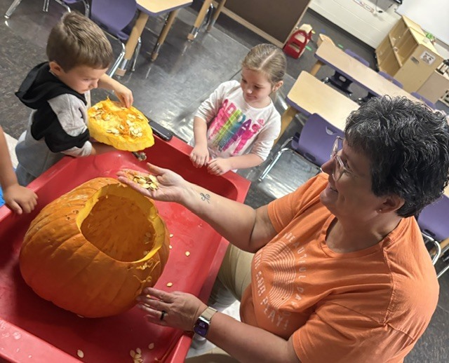 teacher showing seeds to a student while another student explores the top of the pumpkin
