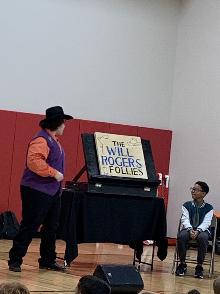 Student and actor on stage with a sign that says The Will Rogers Follies.