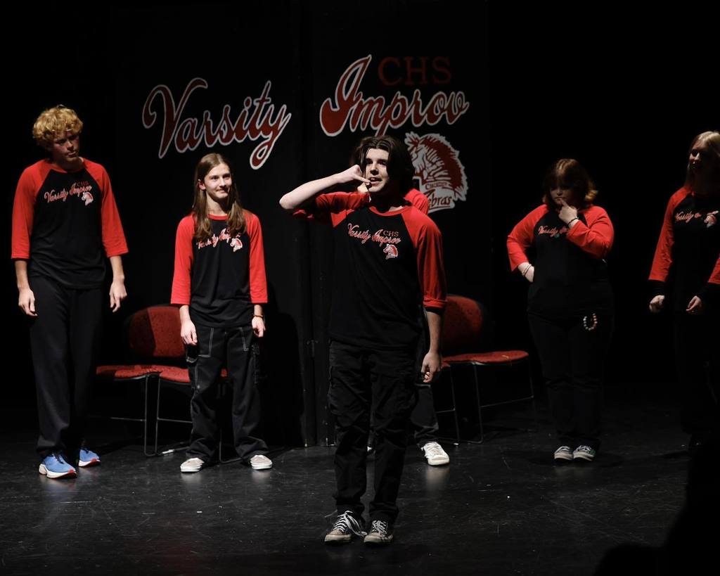 Students in black/red shirts "Improve" on stage micking on a phone