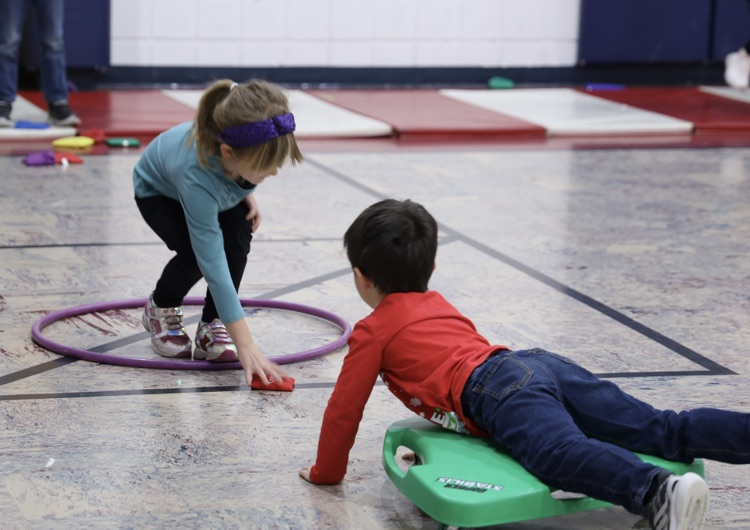Little boy riding a scooter passing a beanbag to a girl