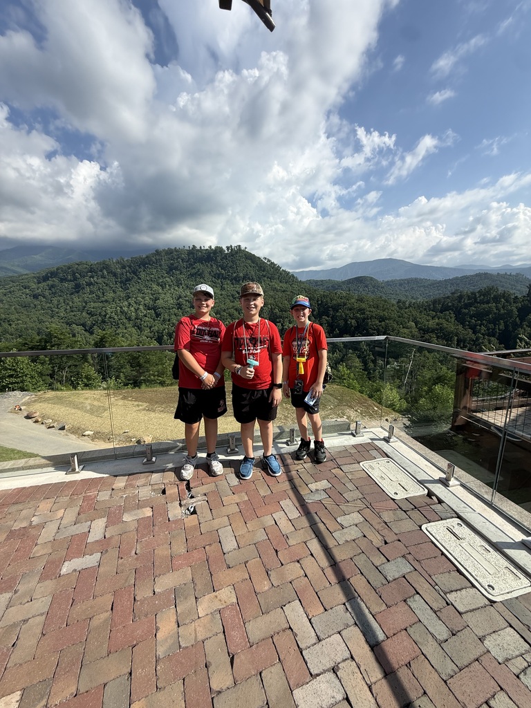 Three Claremore students stand together at a mountain overlook in the Smoky Mountains with green hills and a cloudy blue sky in the background.