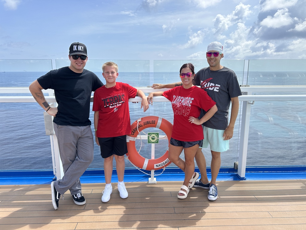 Four family members wearing Claremore Zebra shirts pose on the deck of a cruise ship with the ocean in the background.