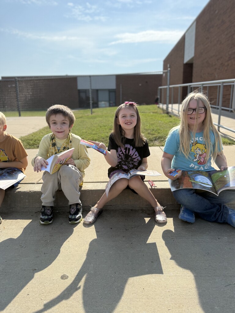 students sitting on sidewalk, smiling and holding books
