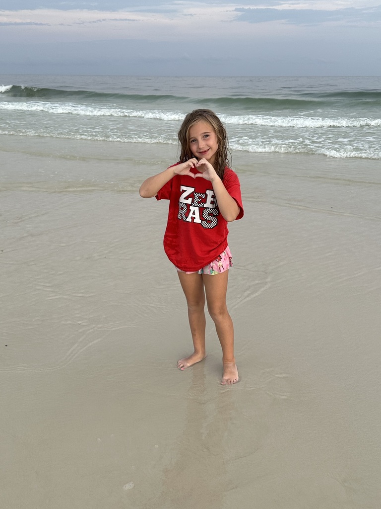 Natalie Stanart stands barefoot on the beach wearing a Claremore Zebras shirt with the ocean and waves in the background.
