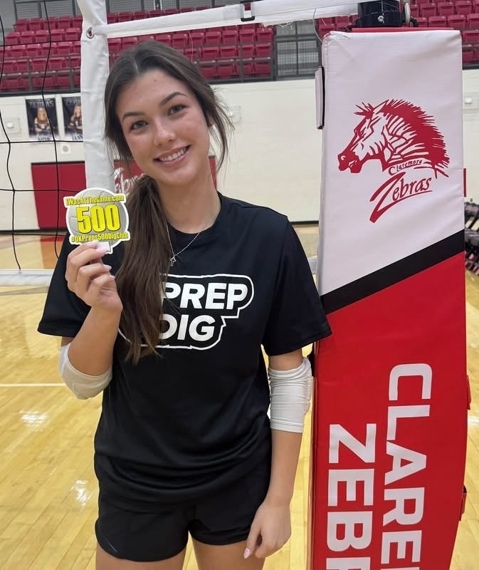 Madison Godding holds her 500 career digs milestone sticker beside the volleyball net.