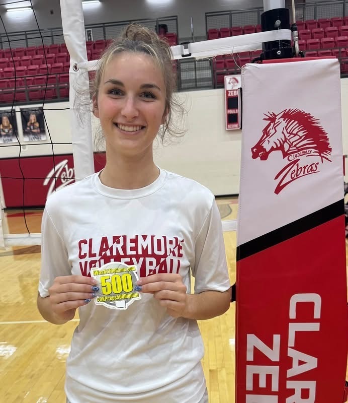 Grace Baughman holds her 500 career digs milestone sticker beside the volleyball net.
