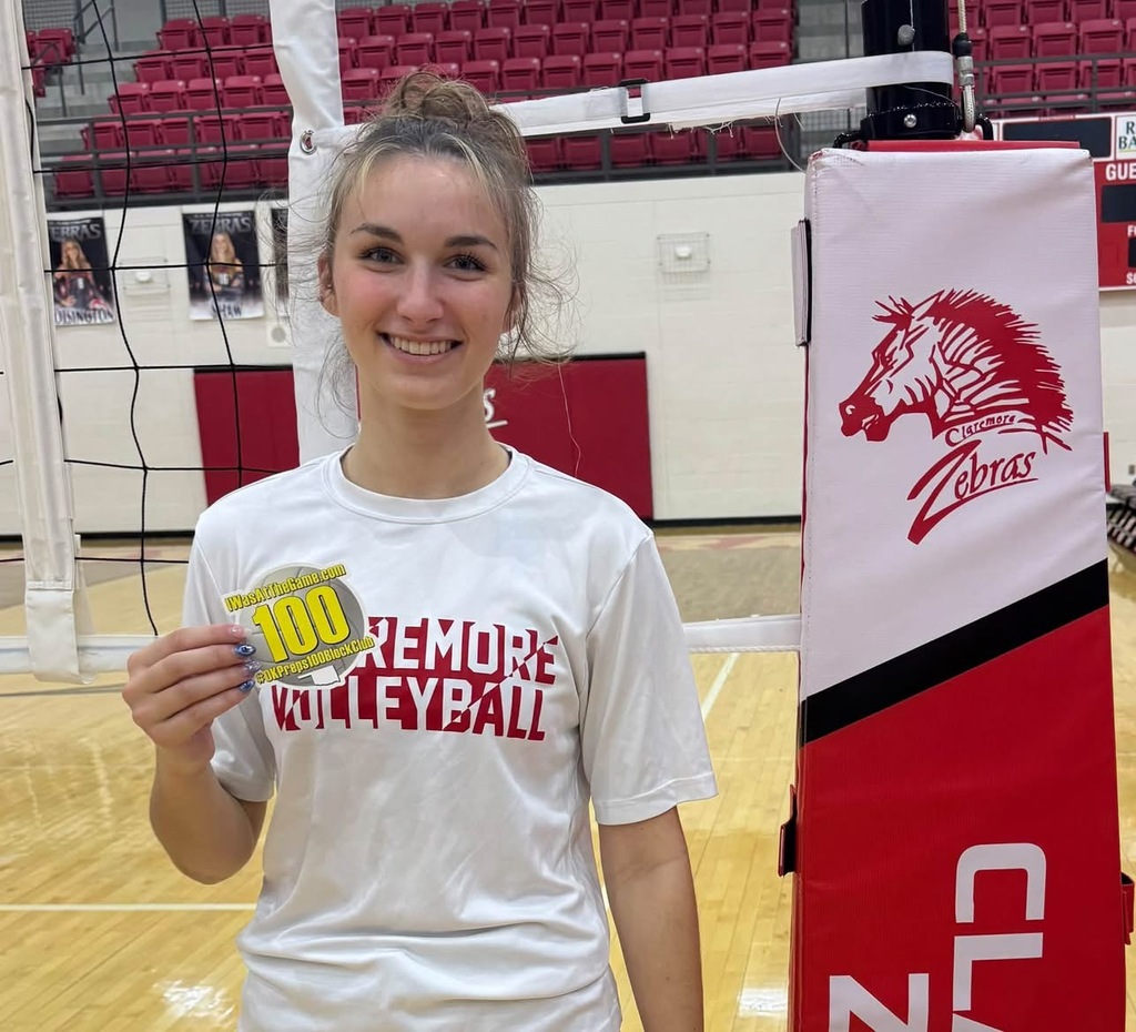 Grace Baughman holds her 100 career blocks milestone sticker beside the volleyball net.