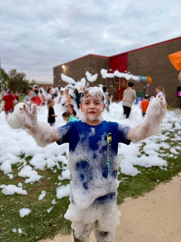 A student covered in foam smiles and holds his hands toward the camera.