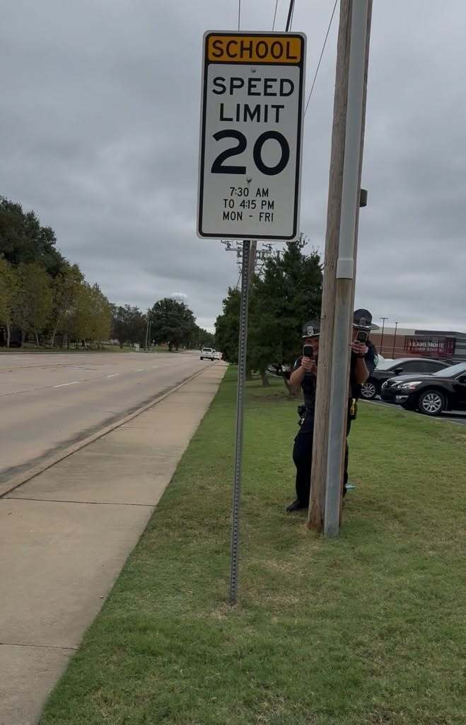 A Claremore SRO hides behind a utility pole near a school zone speed limit sign.