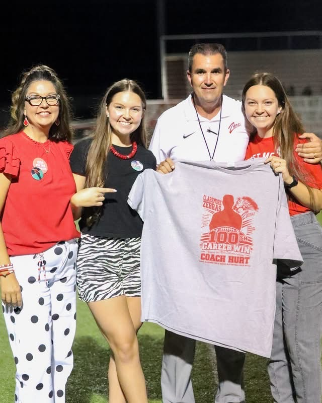 coach Hurd with his wife and two daughters on the field, holding up a 100 wins T-shirt.