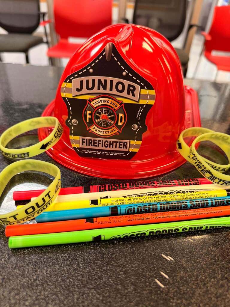 Red junior firefighter helmet surrounded by fire safety bracelets and colorful pencils.