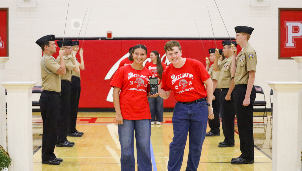 Two students smile and display an item while standing between NJROTC cadets during the Homecoming assembly.
