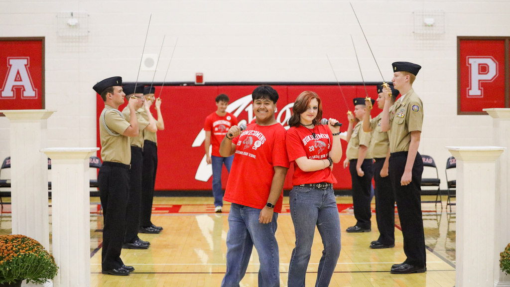 Two students in red Homecoming shirts pose with microphones between rows of NJROTC cadets.