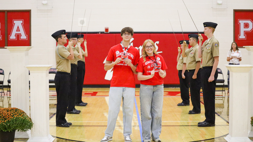 Two students in red Homecoming shirts pose between NJROTC cadets during the assembly.