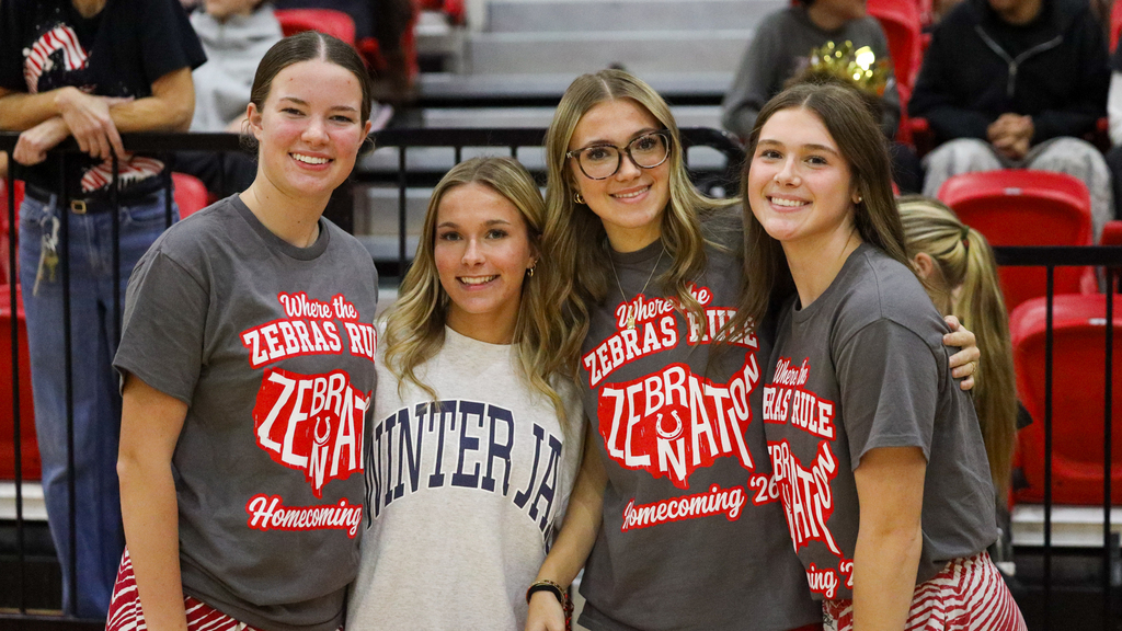 Four students smile together in the gym, three wearing gray Zebra Nation Homecoming shirts.