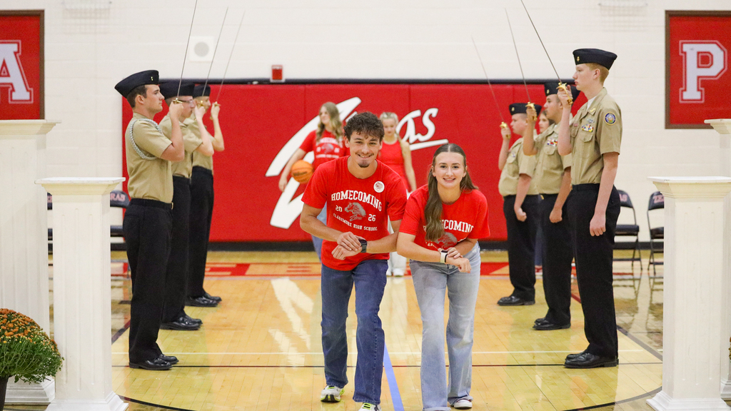 Two students in red Homecoming shirts walk between NJROTC cadets holding raised swords during the assembly.
