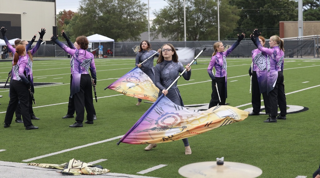 Color guard members perform with colorful clock-themed flags while band members pose behind them.