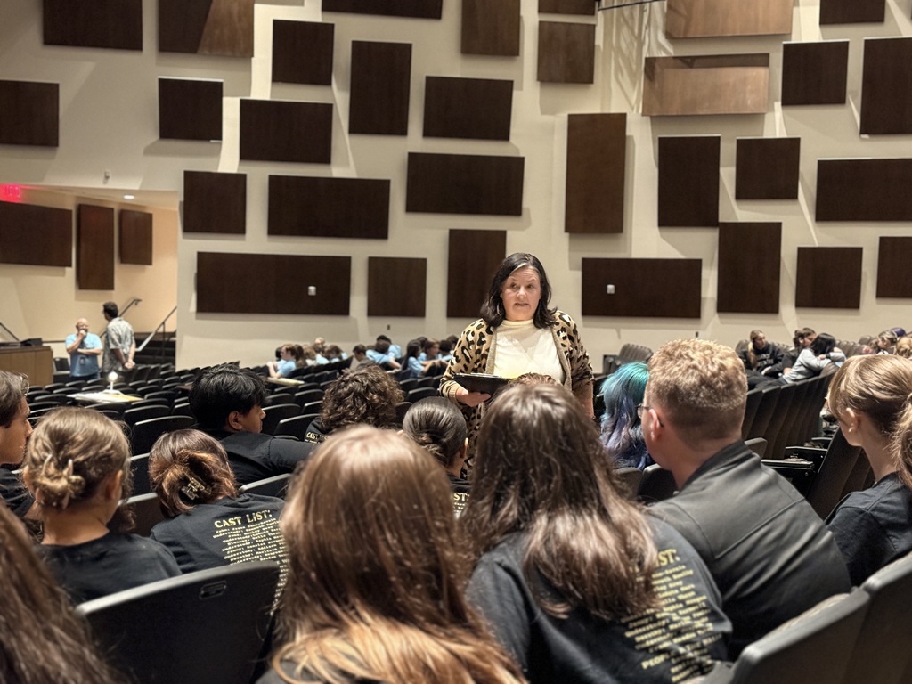 A teacher speaks with drama students seated in the auditorium.