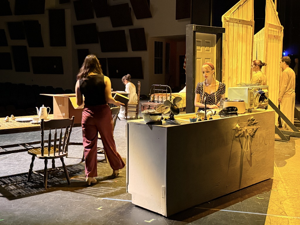 Cast members perform on a detailed stage set featuring a bedroom and kitchen.