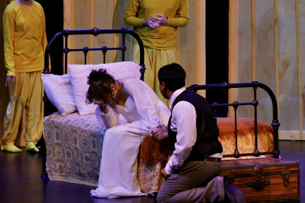 Two student actors perform an emotional scene beside a bed on stage.