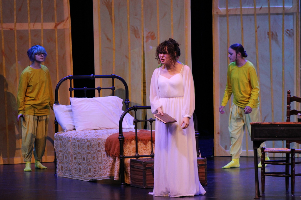 A student actor in a white dress performs alongside two actors in yellow costumes.