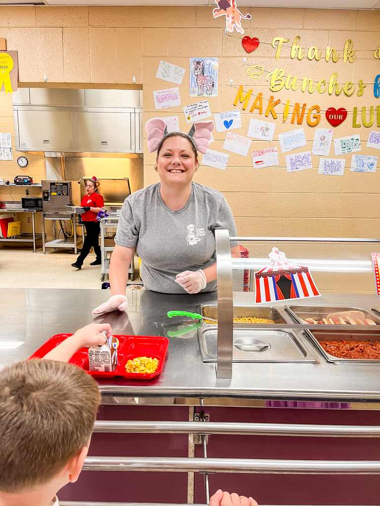 A smiling Claremont cafeteria employee wearing elephant ears serves lunch to a student.