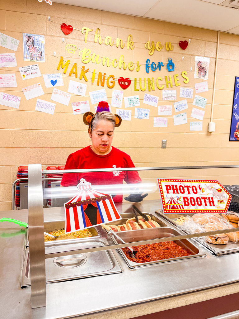 A cafeteria employee wearing a monkey headband prepares lunch behind a circus-decorated serving counter.
