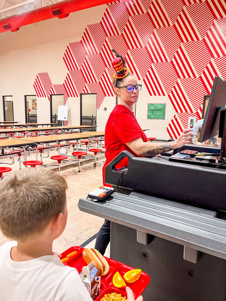 A cafeteria employee wearing a circus ringmaster hat helps a student at checkout.