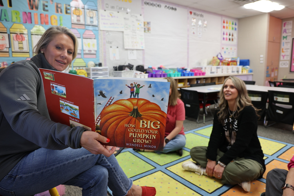 A principal reads a fall-themed book, How Big Could Your Pumpkin Grow?, to teachers gathered on a colorful classroom rug as they get ready for fall break.
