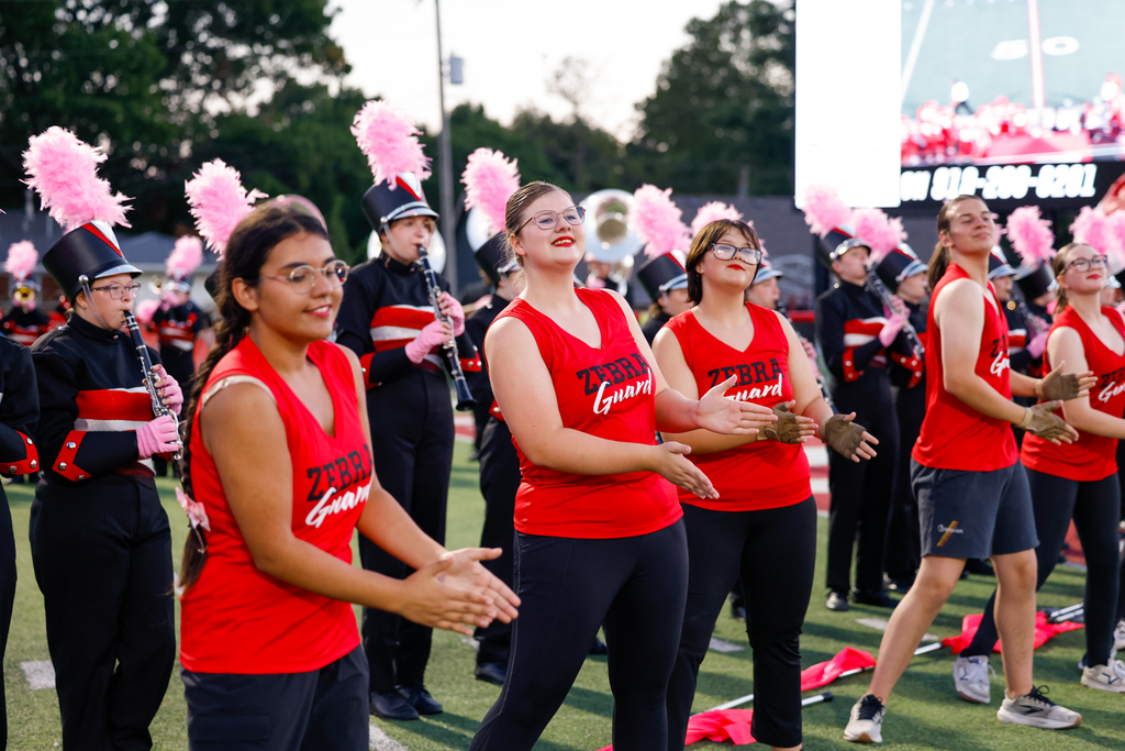 Zebra Guard members in red shirts perform alongside the marching band wearing pink plumes.