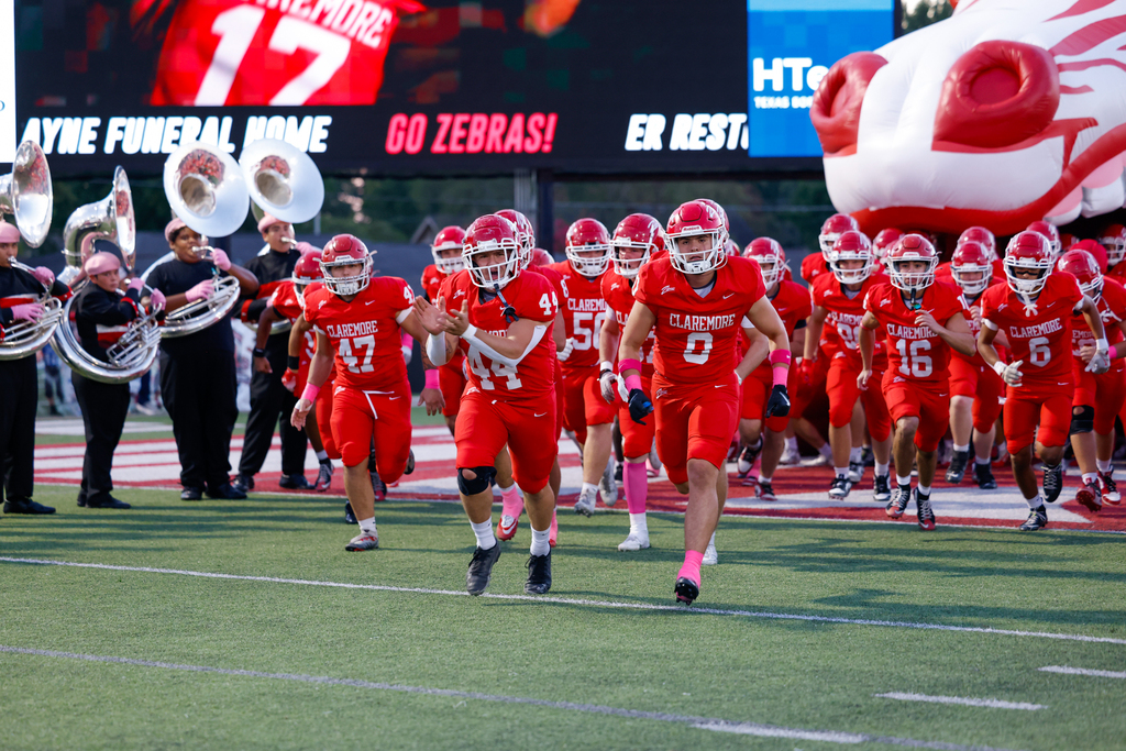 Claremore football players run onto the field in red uniforms with pink accessories for Pink Out Night.
