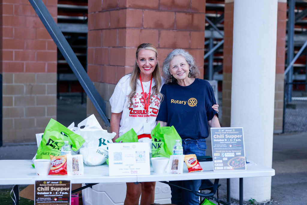 Two women representing the Claremore Public School Foundation and Rotary Club smile behind a table with chili supper meals, sponsor bags, and event information.