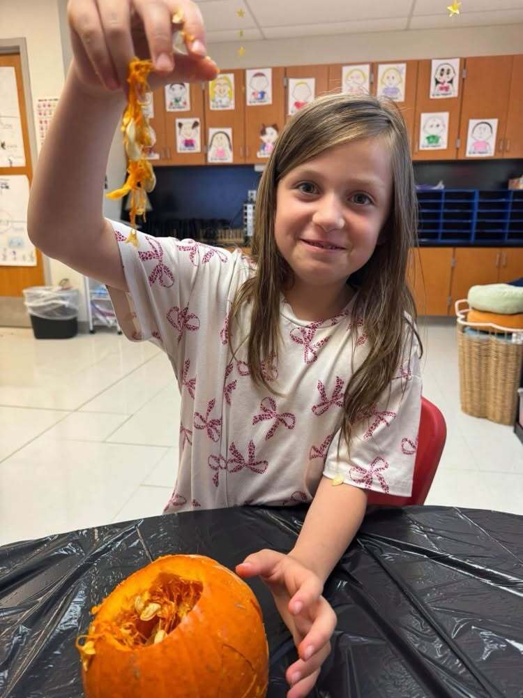 A student smiles while holding up a handful of pumpkin seeds and pulp over a carved pumpkin.