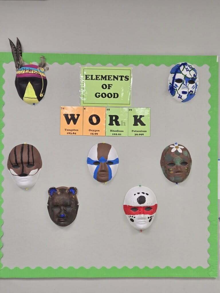 Seven hand-painted student masks displayed on a bulletin board with a green border and an “Elements of Good Work” sign.