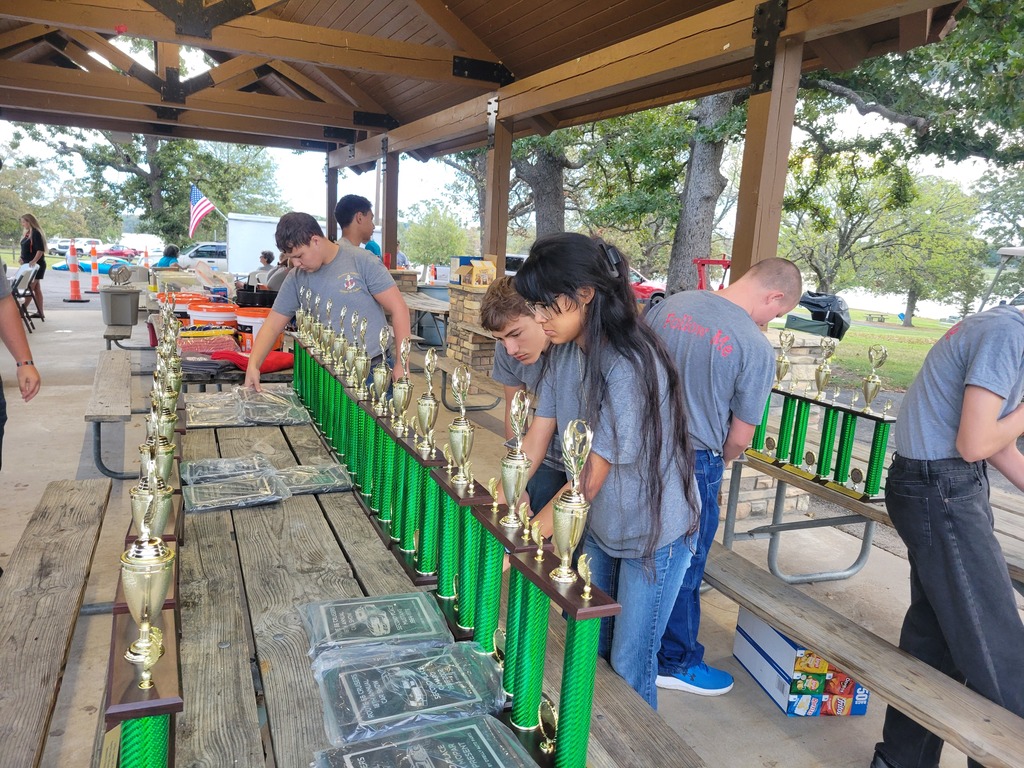 NJROTC cadets arrange large green and gold trophies on picnic tables under a pavilion.