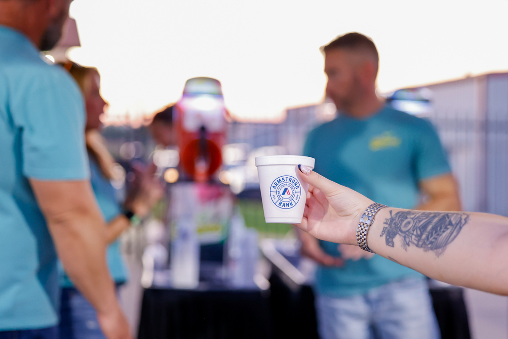 A hand holds an Armstrong Bank cup in front of the Blended Margarita Co. slushie stand.