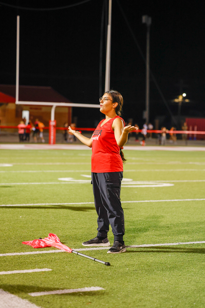 A color guard member gestures with both hands while standing beside a red flag on the football field.
