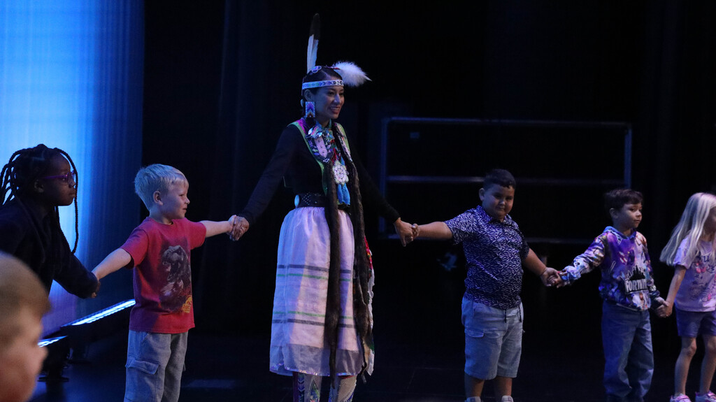 Rising Buffalo dancer holding hands with elementary students on stage.