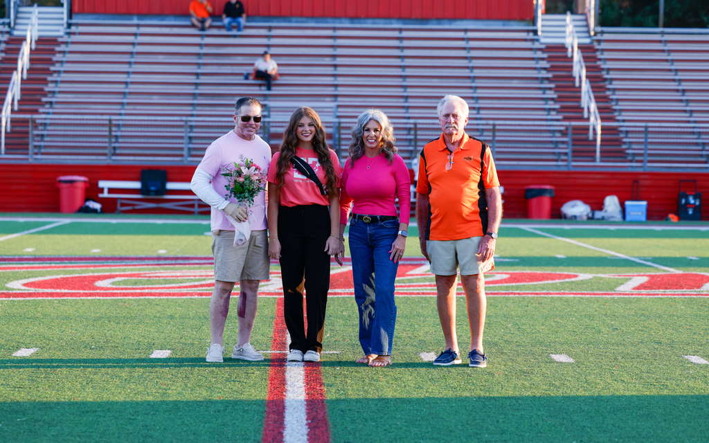 Senior student athletic training aid wearing a Claremore shirt poses with three family members at midfield.