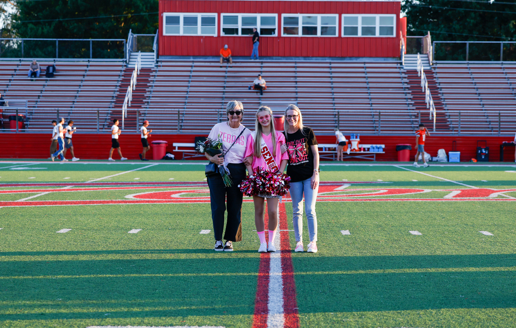 Senior dance member in a pink outfit and senior sash holds pom-poms while posing with two family members.