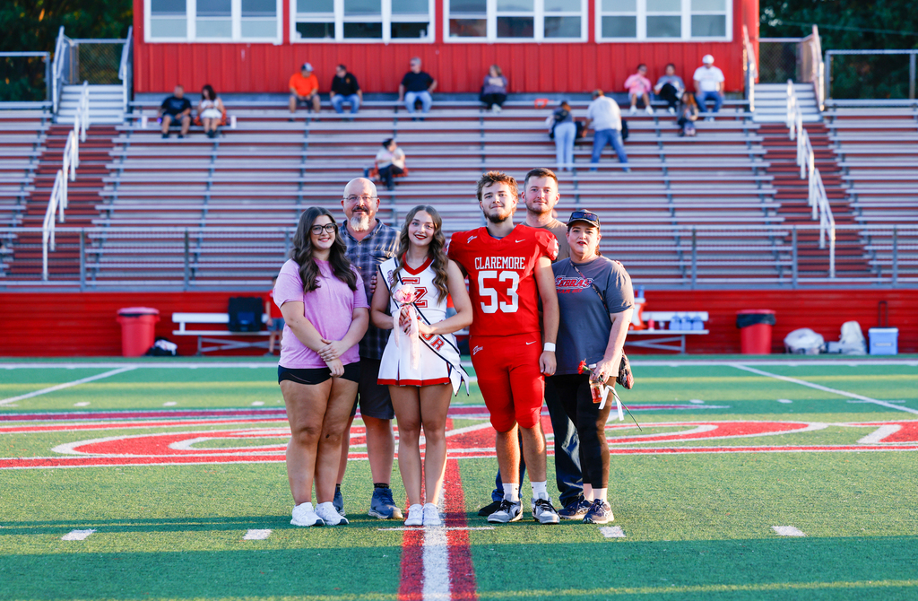 Claremore football player #53 and a cheerleader seniors pose with family members on the football field.