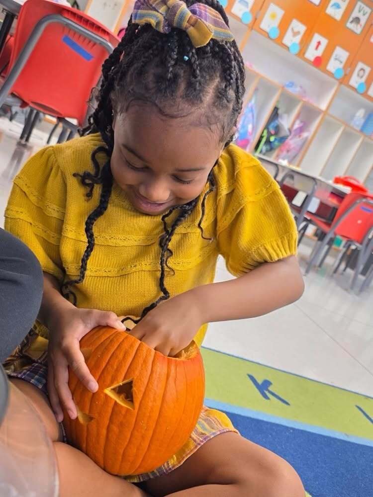 A smiling student in a yellow shirt reaches inside a carved pumpkin.