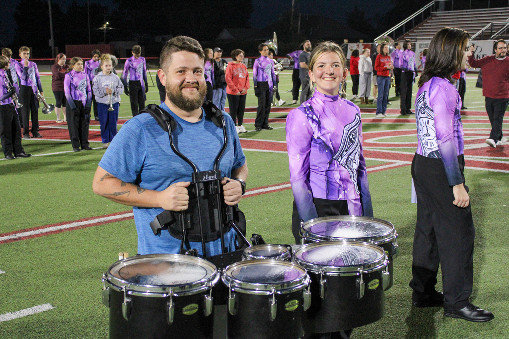 A parent wearing a drum harness poses with a smiling band student.