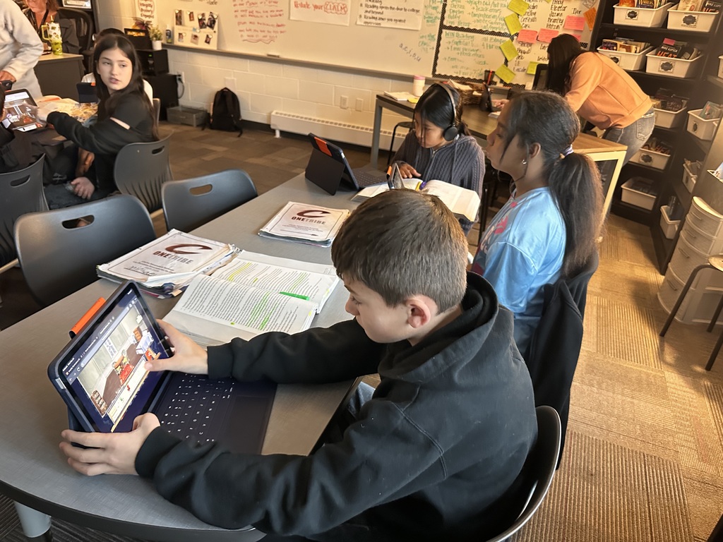 A group of sixth-grade students sit at a table in a language arts classroom. Several students are using iPads to arrange items in a digital room scene while their open books and highlighted text lie on the table. Other students read or work independently in the background.