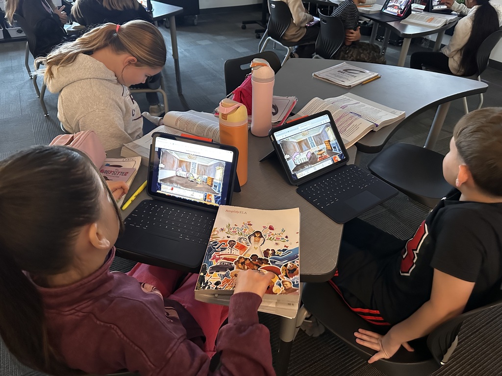 Two students work at a table with open books and iPads showing a digital illustration of a bedroom scene from a story. One student points at the screen while the other reads. Additional groups of students are working at nearby tables in the classroom.