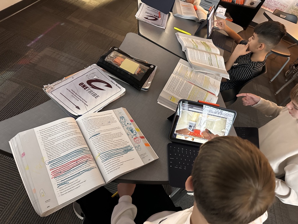 Students sit around a table with open ELA textbooks covered in colorful highlighting. Each student is using an iPad to place furniture inside a digital layout of a story setting. Notebooks and “One Tribe” folders are also visible on the table.