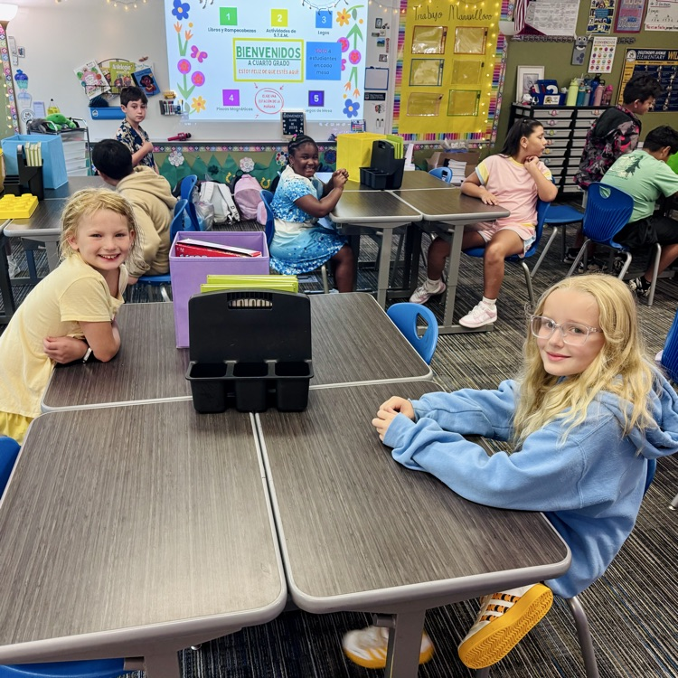 two girls sit at desks in front of a whiteboard
