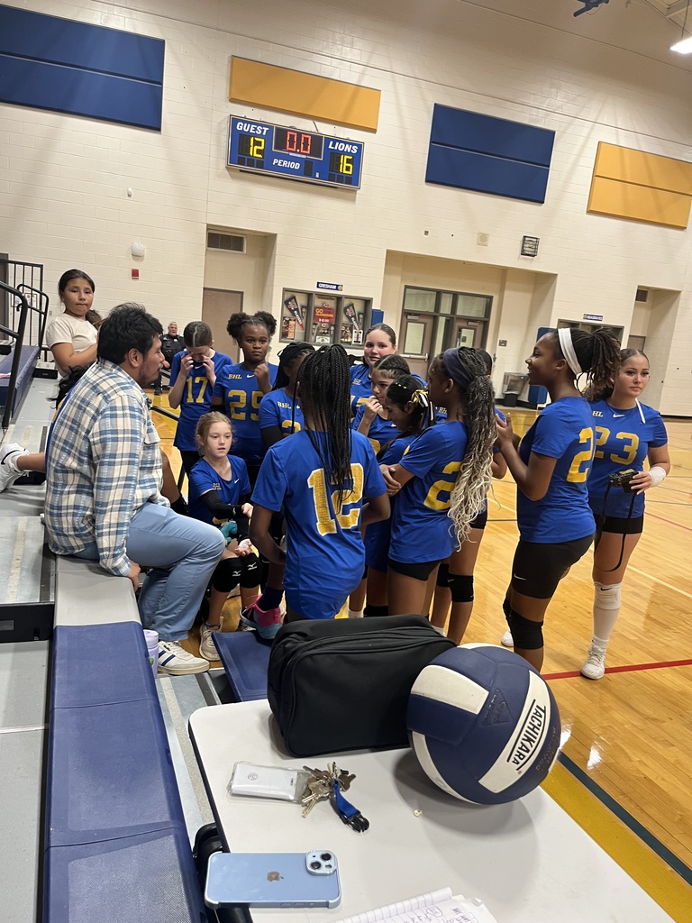 A volleyball team in blue and gold jerseys gathers together on a basketball court next to their coach sitting on the bleachers, with a volleyball resting in the foreground.